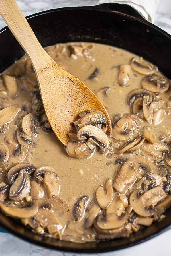 Sliced mushrooms and sauce simmering in cast iron skillet with wooden spoon.
