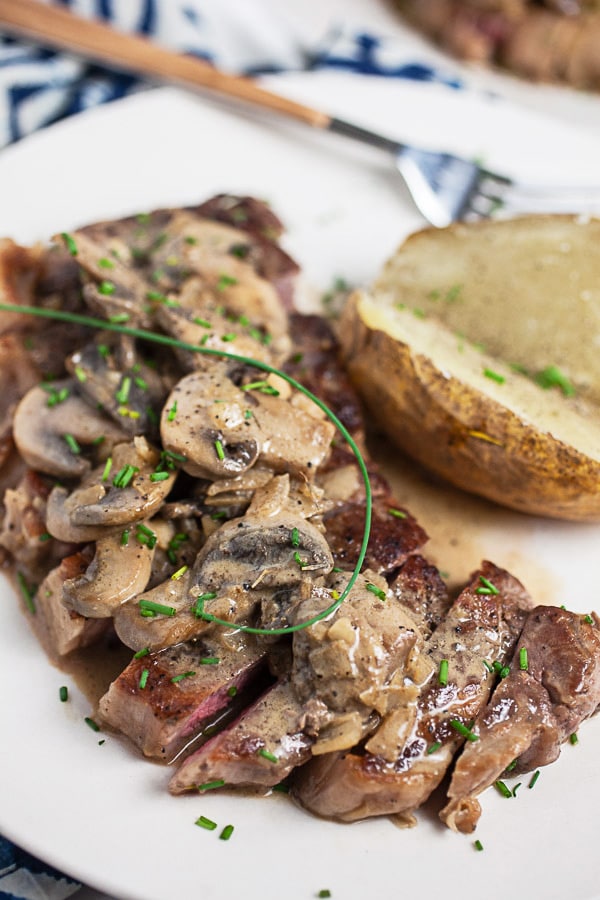 Sliced steak and mushrooms garnished with fresh chives on white plate with baked potato.
