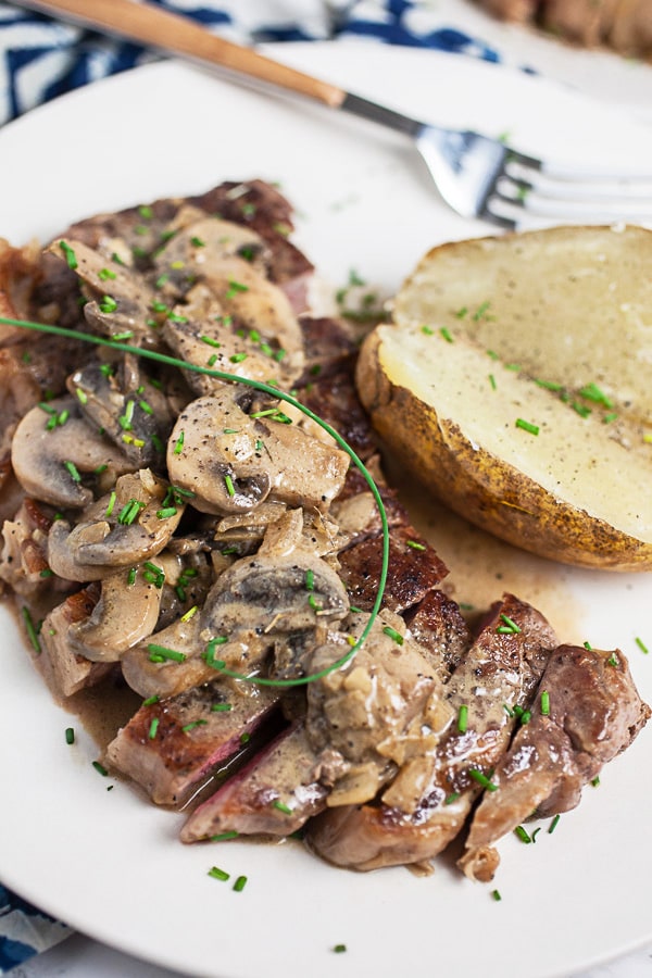 Sliced Steak Diane and mushrooms on white plate with baked potato.