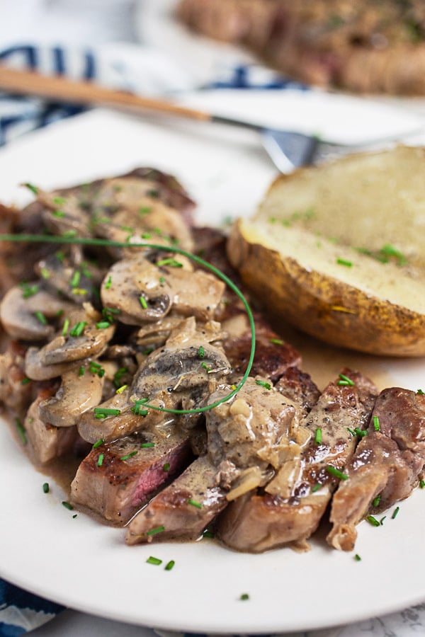 Sliced Steak Diane with mushroom sauce on white plate with baked potato.