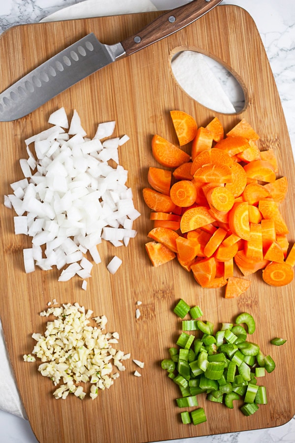 Minced garlic, onions, celery, and carrots on wooden cutting board with knife.