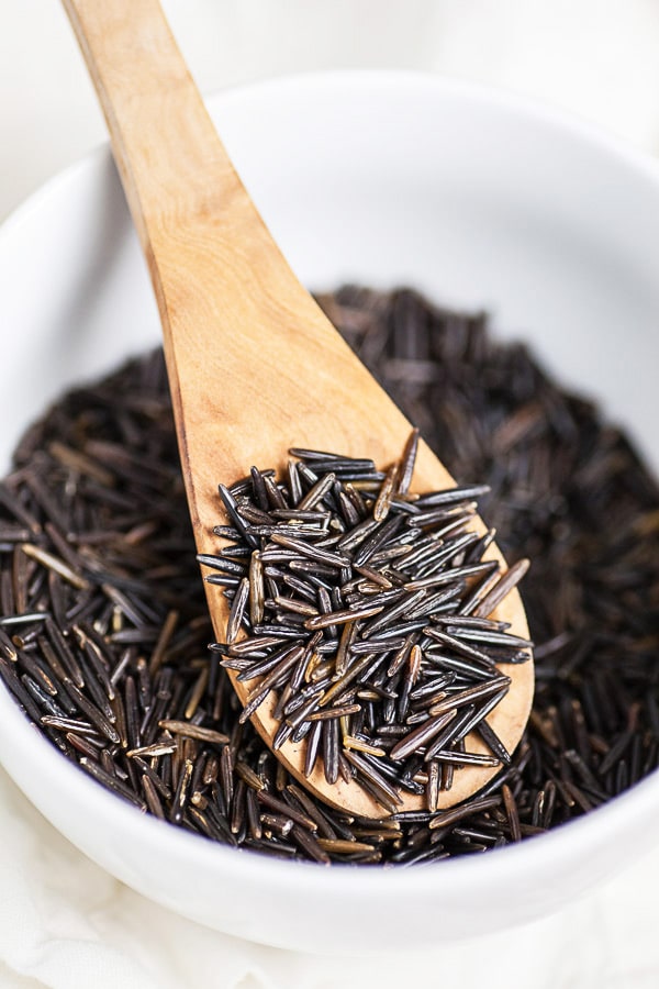 Uncooked wild rice lifted from white bowl on wooden spoon.