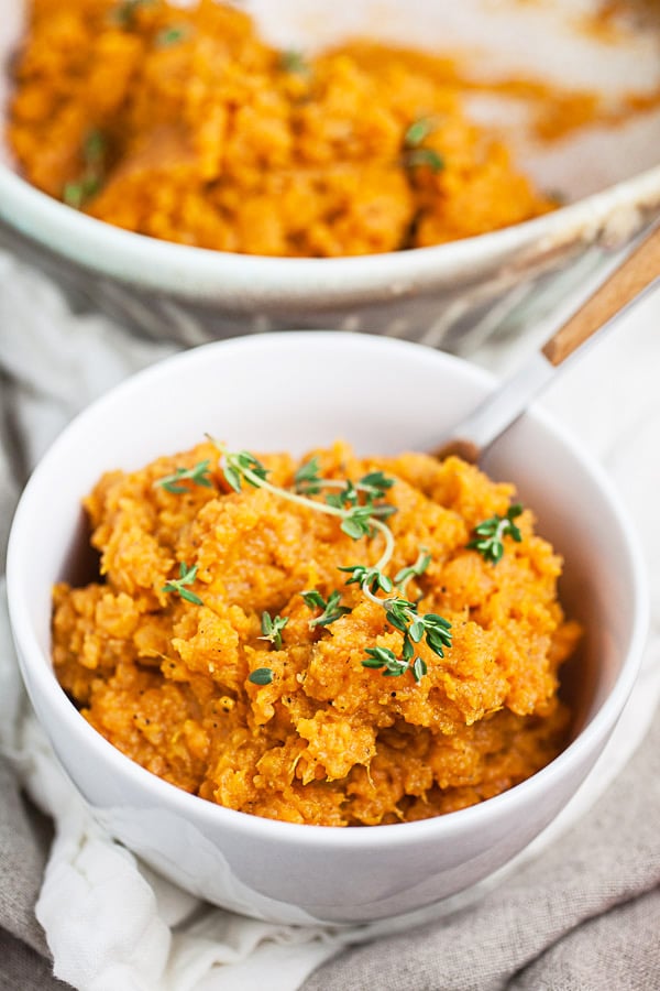 Mashed sweet potatoes in small white bowl in front of ceramic bowl.