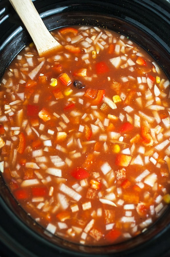 Uncooked broth and soup ingredients in slow cooker with wooden spoon.