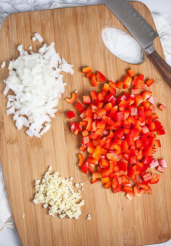 Minced garlic, onions, and red bell peppers on wooden cutting board with knife.