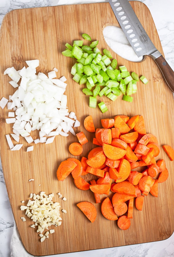 Minced garlic, onions, celery, and chopped carrots on wooden cutting board with knife.