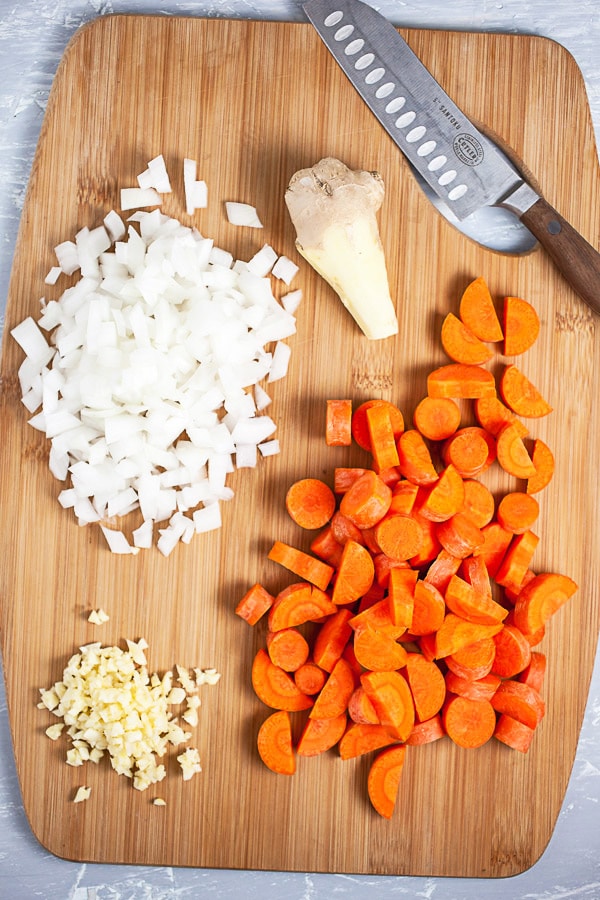 Minced garlic, onions, carrots, and ginger root on wooden cutting board with knife.