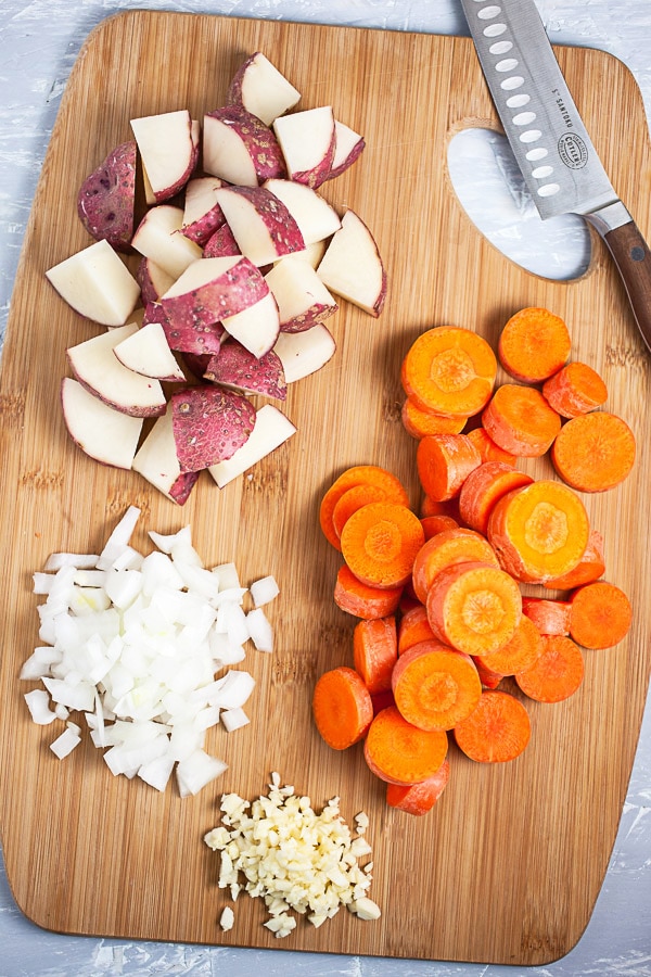 Minced garlic and onions and chopped red potatoes and carrots on wooden cutting board with knife.