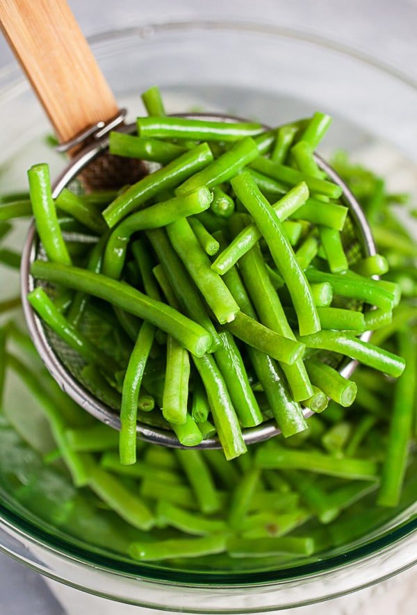 Blanched green beans on strainer lifted from glass bowl of ice water.