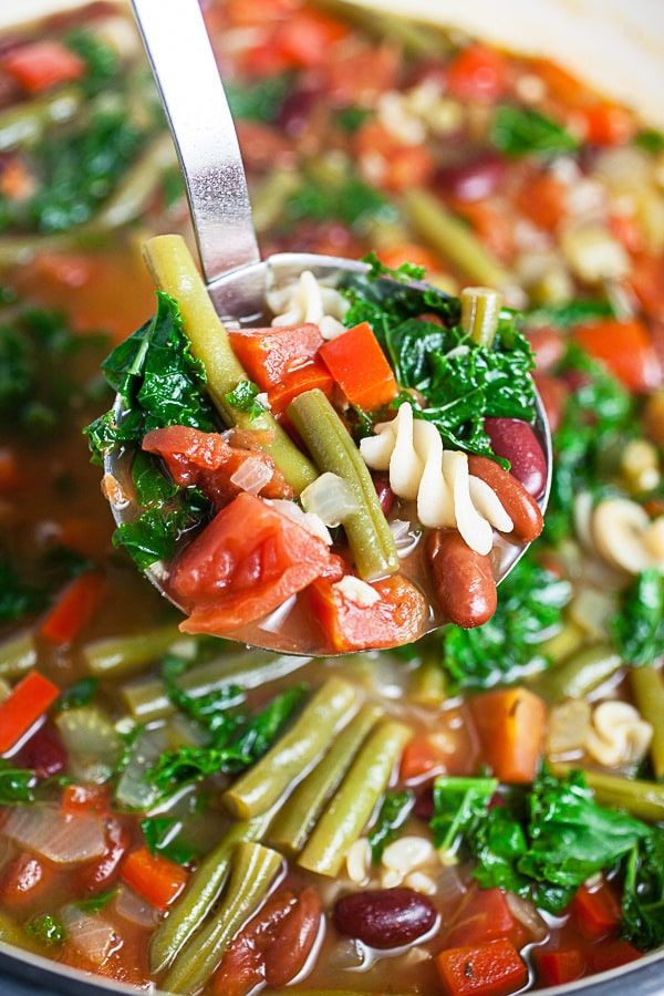 Scoop of vegetable noodle soup lifted from Dutch oven in metal ladle.