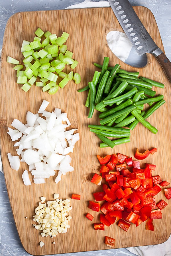 Minced garlic, onions, celery, red bell peppers, and green beans on wooden cutting board with knife.