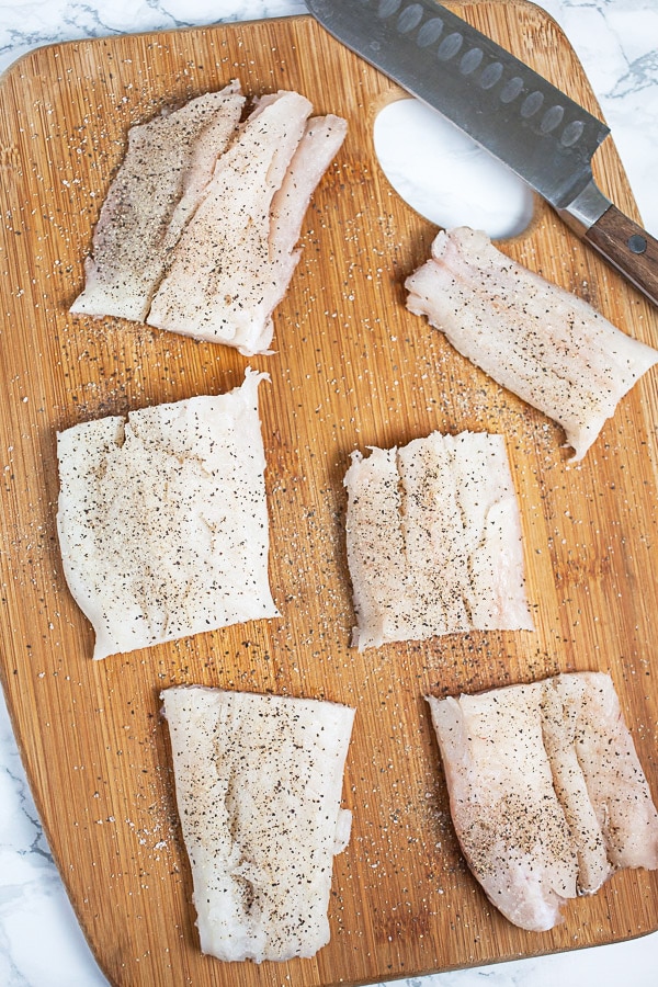 Raw fish fillets sprinkled with salt and pepper on wooden cutting board with knife.