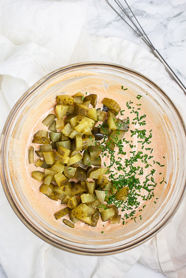 Dressing with chopped dill pickles and minced chives in small glass bowl next to whisk.