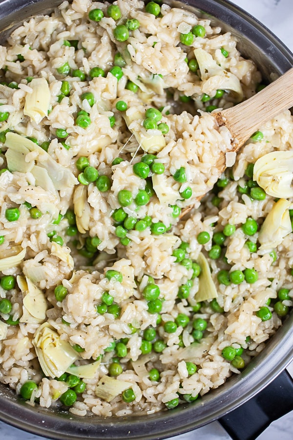 Cooked risotto with peas, artichokes, lemon, and Parmesan cheese in skillet with wooden spoon.