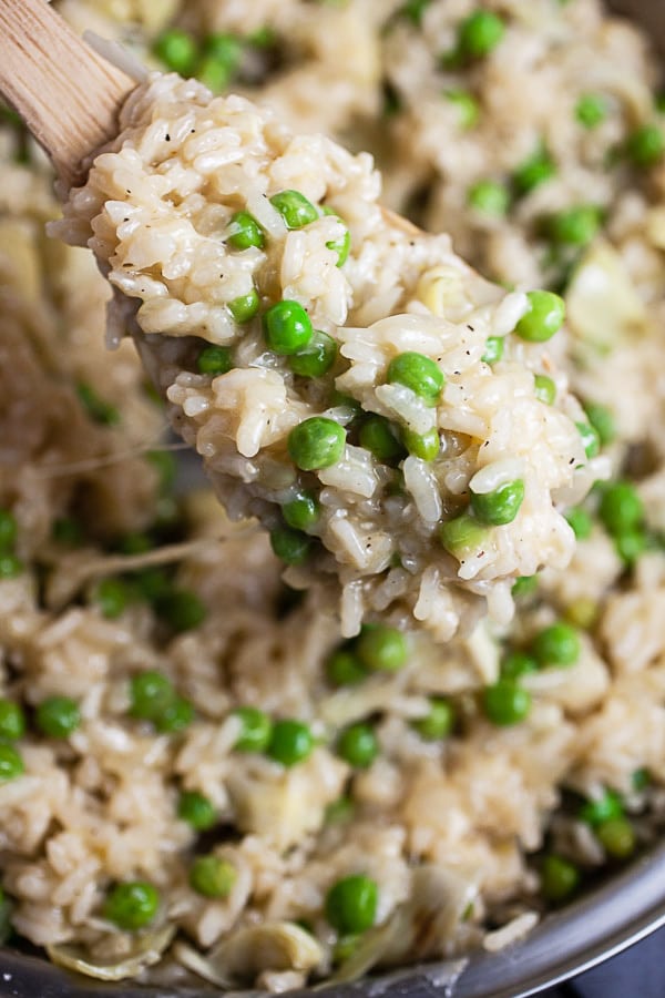 Scoop of cooked cheesy Arborio rice with peas lifted from skillet on wooden spoon.