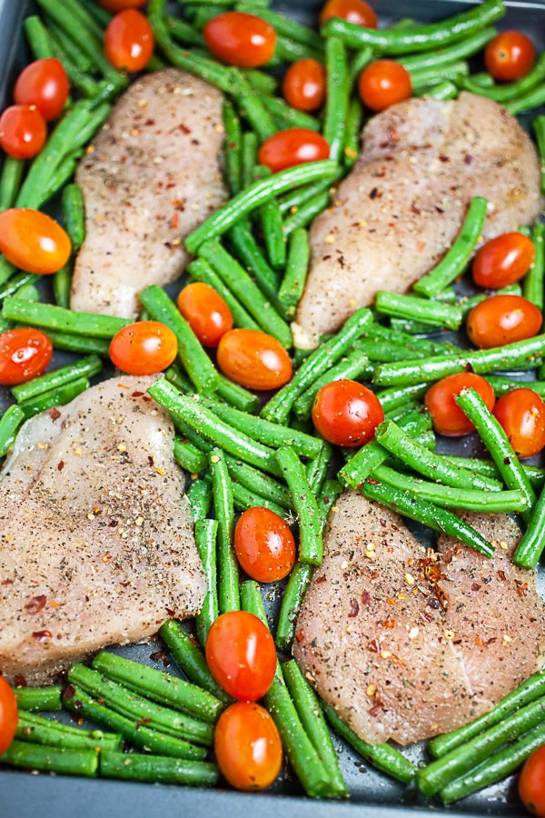 Uncooked chicken breasts, green beans, and tomatoes on metal baking sheet.