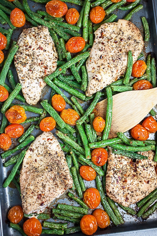 Cooked chicken breasts, green beans, and tomatoes on metal baking sheet with wooden spatula.