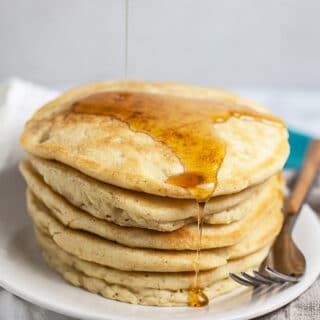 Maple syrup pouring onto stack of gluten free pancakes on white plate.