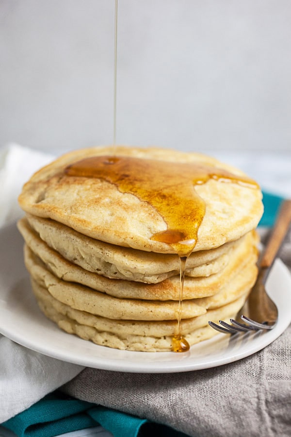 Maple syrup pouring onto stack of old fashioned pancakes on white plate.