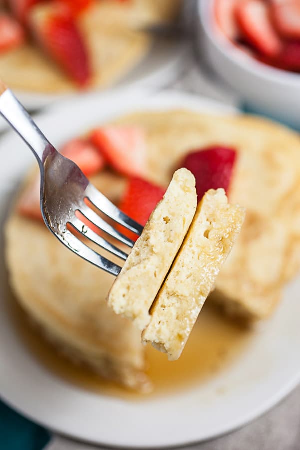 Forkful of pancakes lifted from plate of pancakes topped with fresh strawberries.