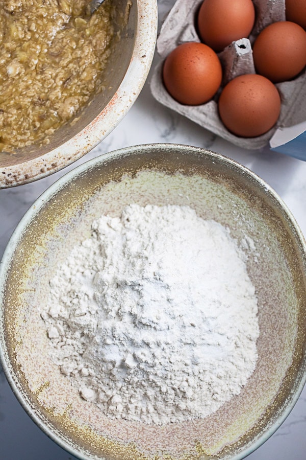 Dry and wet ingredients in two separate ceramic bowls next to carton of eggs.