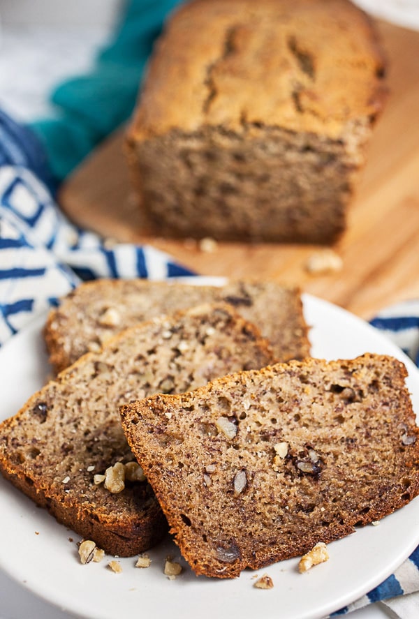 Sliced banana bread on small white plate next to loaf on wooden cutting board.