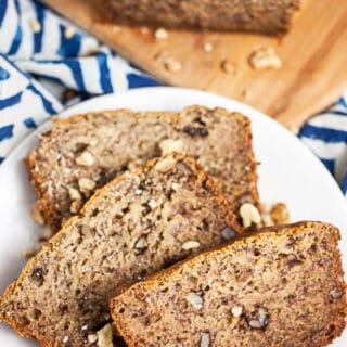 Roasted banana bread slices on small white plate next to loaf on wooden cutting board.