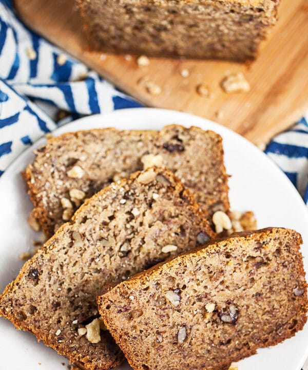 Roasted banana bread slices on small white plate next to loaf on wooden cutting board.