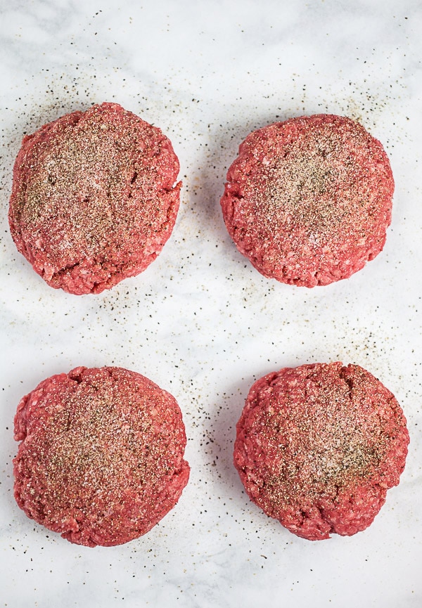 Raw ground beef patties seasoned with garlic powder, onion powder, salt and pepper on white surface.