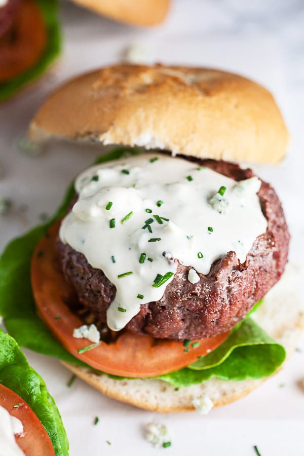 Grilled burgers with lettuce, tomato, and blue cheese sauce on white surface.