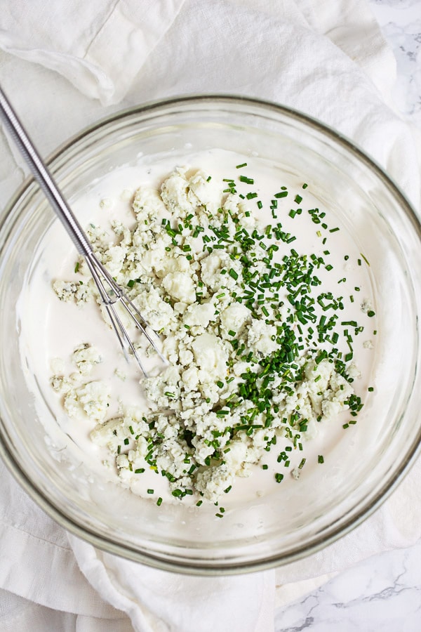 Creamy sauce with blue cheese crumbles and minced chives in small glass bowl with whisk.
