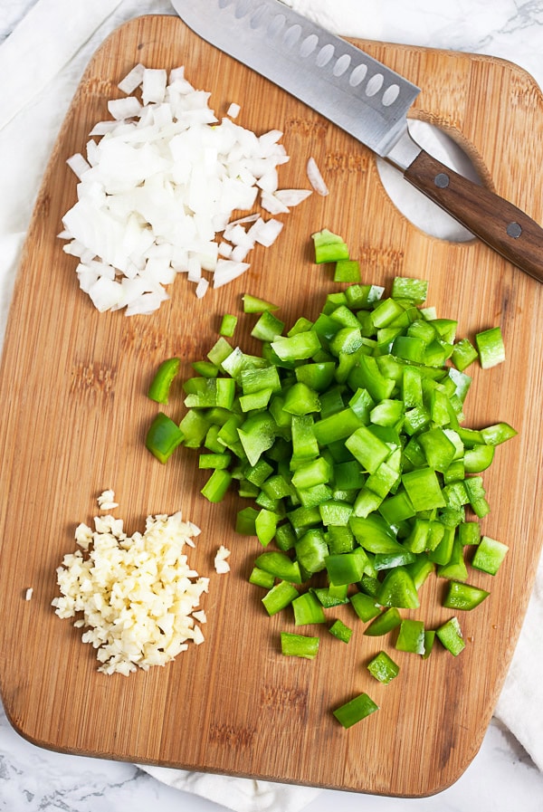 Minced garlic, onions, and green bell peppers on wooden cutting board with knife.