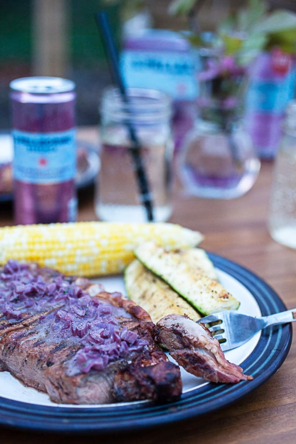 Grilled ribeye steak, zucchini, and corn on the cob next to sparkling water on outdoor table.