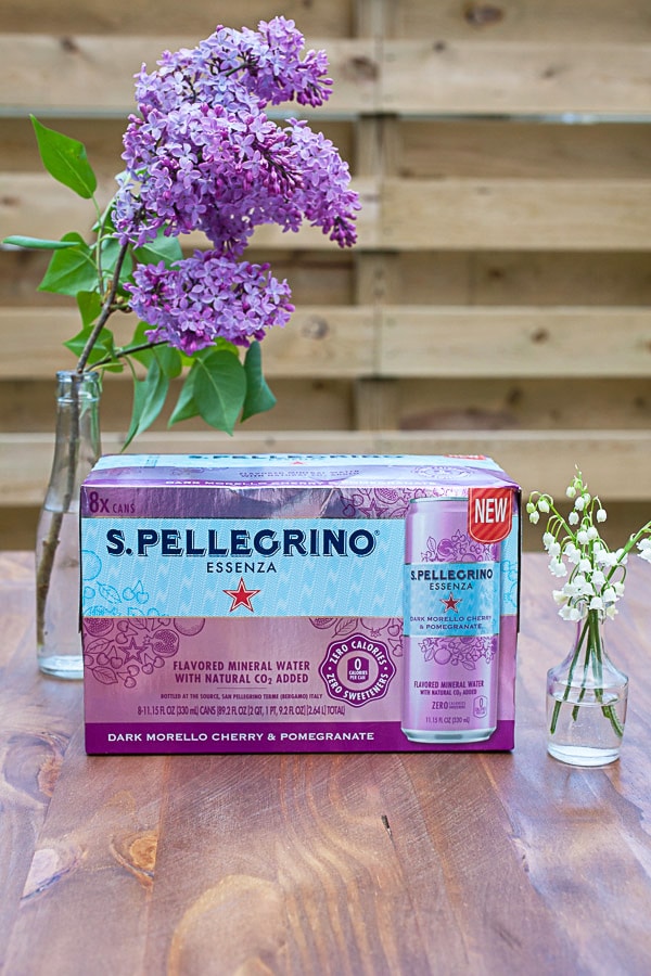 Case of sparkling water on outdoor wooden table next to vase of flowers.