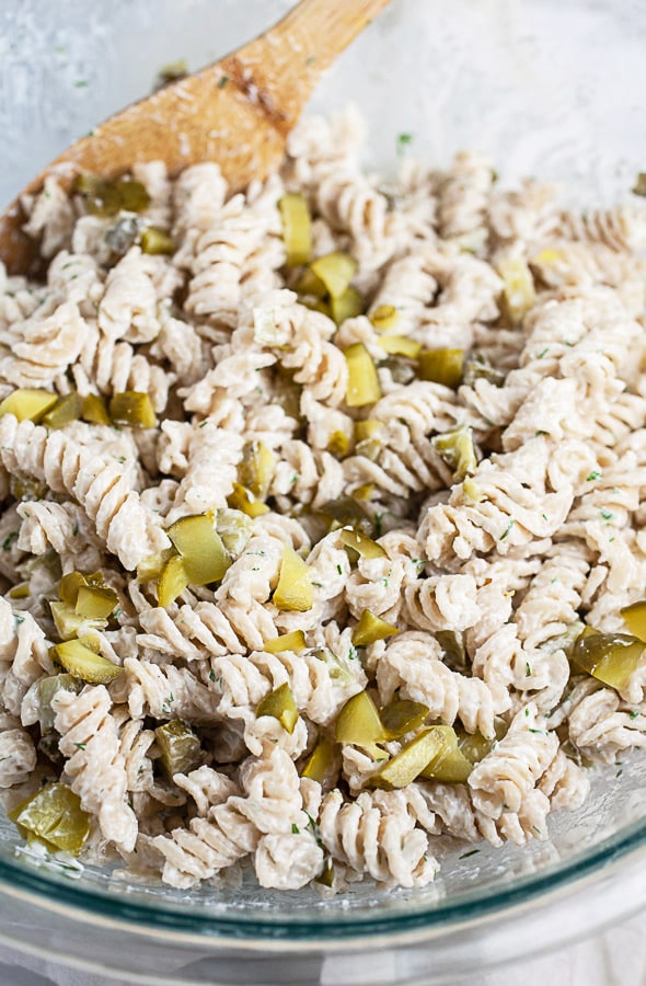 Cooked rotini pasta tossed with dressing and chopped dill pickles in large glass bowl.