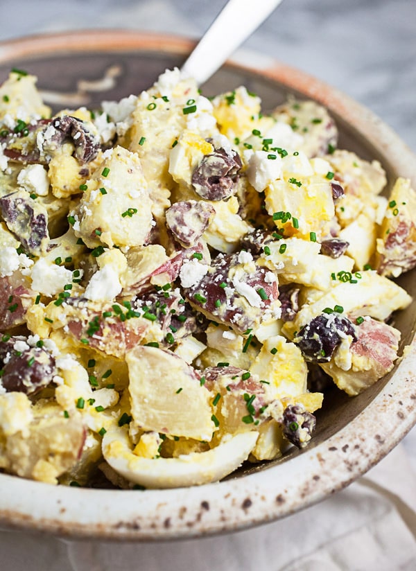 Mediterranean potato salad garnished with chives in ceramic bowl with metal spoon.