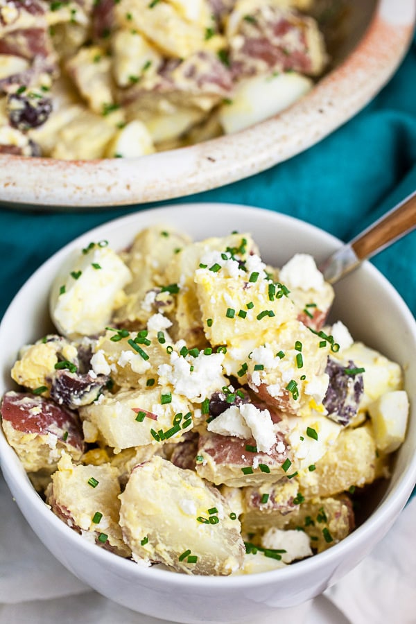 Potato salad with feta in small white bowl next to larger ceramic bowl.