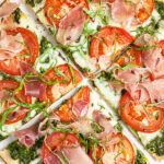 Arugula pesto pizza with tomatoes and prosciutto cut into slices.