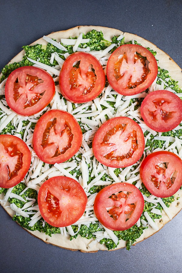 Uncooked pizza crust topped with arugula pesto, sliced tomatoes, and shredded mozzarella cheese.