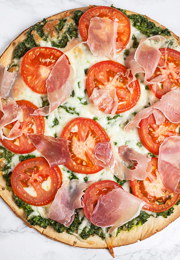 Baked pizza with arugula pesto, sliced tomatoes, mozzarella cheese, and sliced prosciutto.