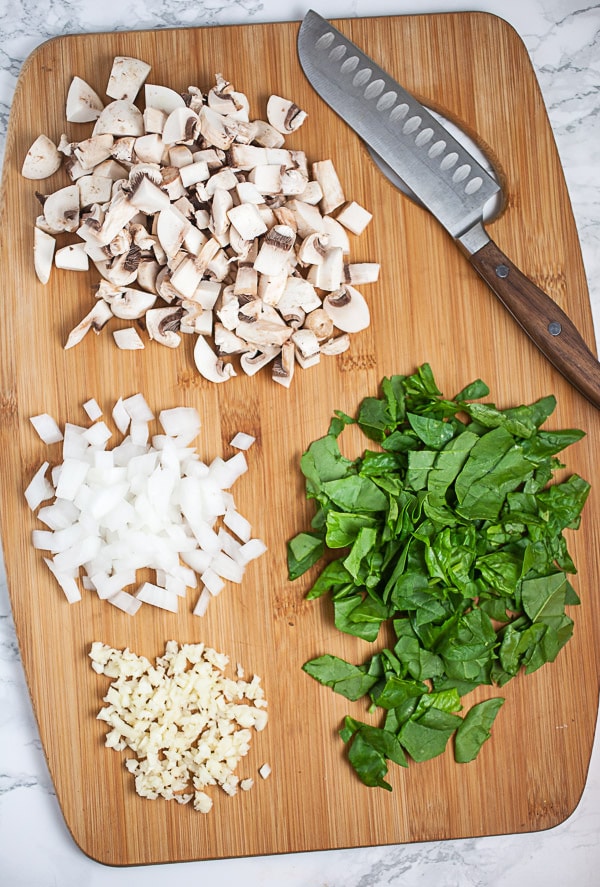 Minced garlic, onions, mushrooms, and spinach on wooden cutting board with knife.