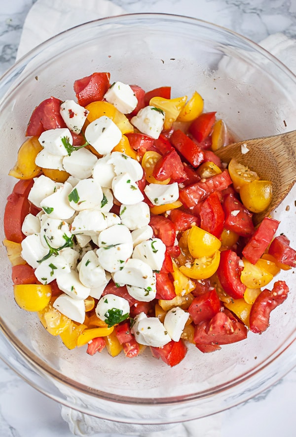 Diced red and yellow tomatoes mixed with dressing topped with fresh mozzarella balls in glass mixing bowl.