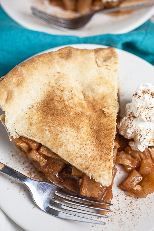 Piece of apple pie with whipped cream and cinnamon on small white plate with fork.