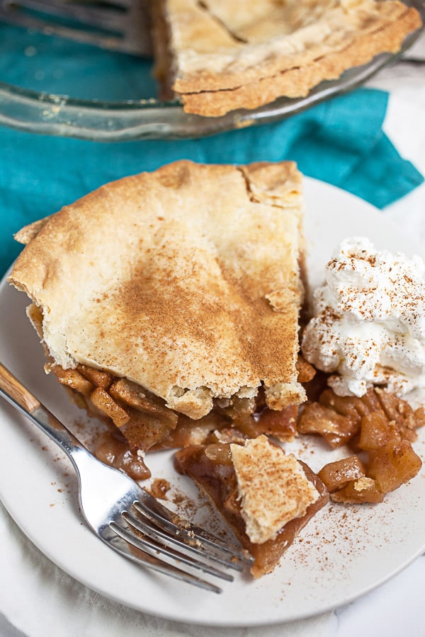 Piece of apple pie cut into with fork on small white plate with whipped cream.