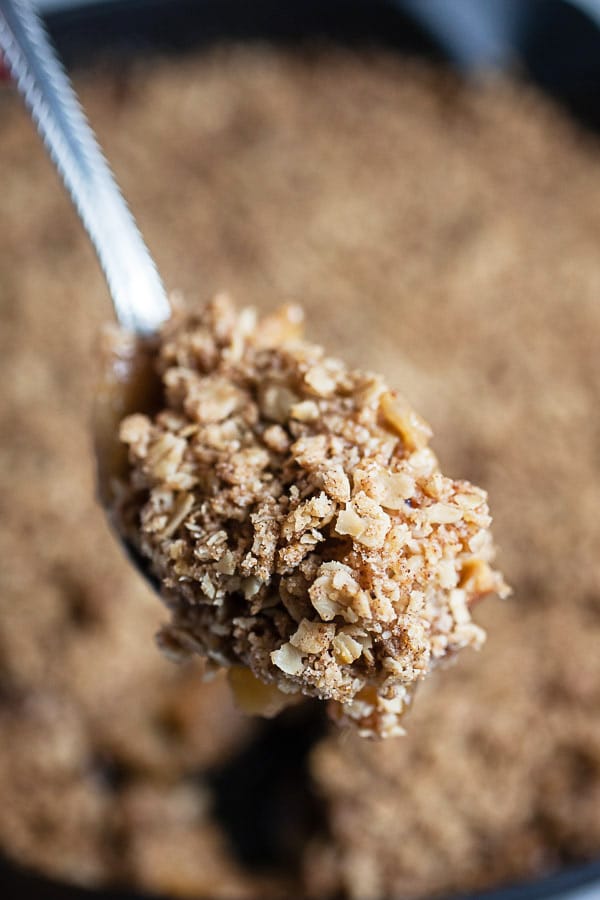 Scoop of baked streusel apple crisp lifted from cast iron skillet on spoon.