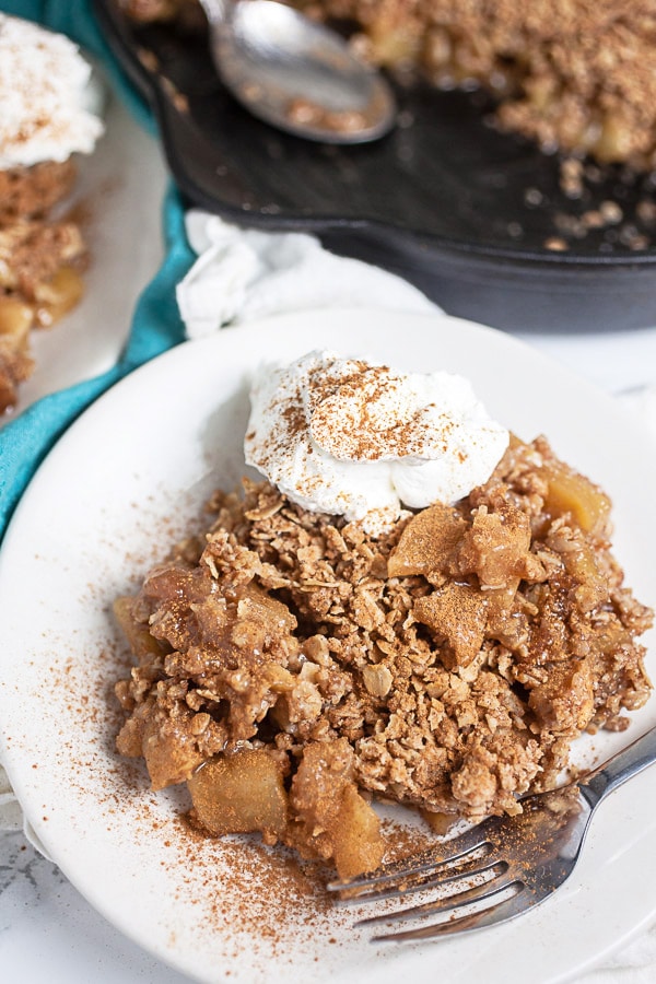 Apple crisp with whipped cream and cinnamon on small white plate with fork.