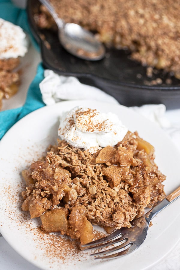 Baked apple crisp with whipped cream and cinnamon on white plate next to cast iron skillet.