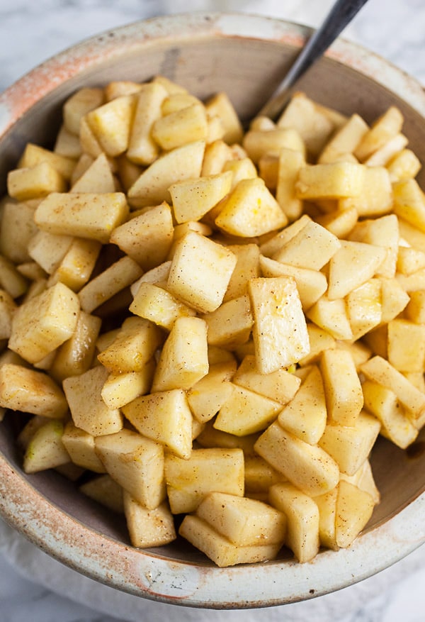 Diced apples tossed with lemon juice and cinnamon in ceramic bowl with spoon.