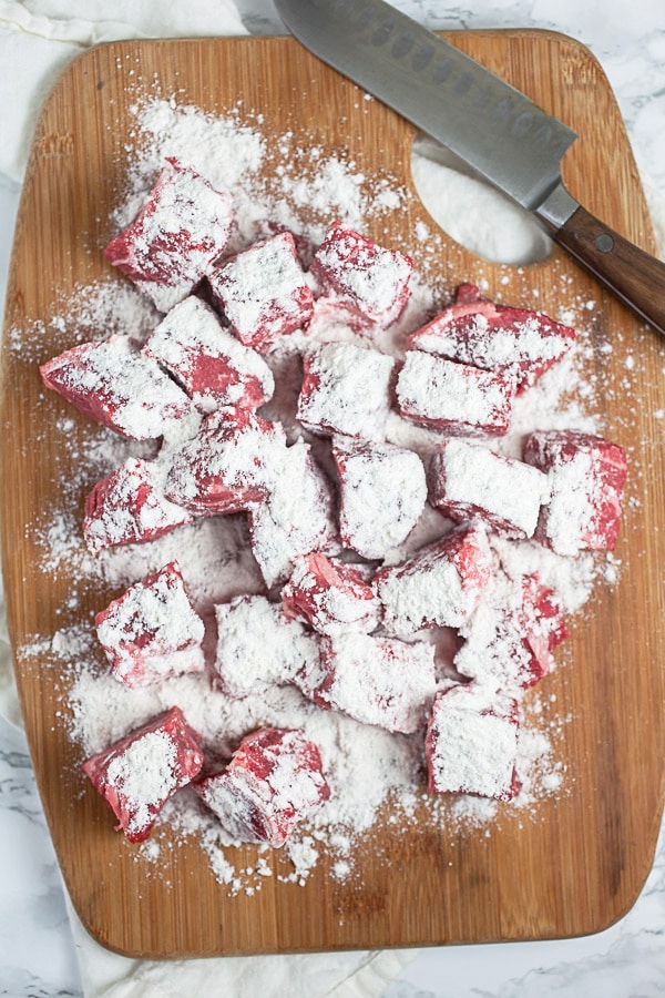 Chunks of raw beef chuck roast dredged in flour on wooden cutting board.