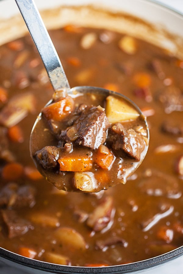 Scoop of cooked beef stew lifted from Dutch oven in metal ladle.